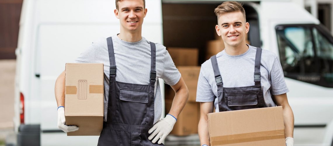 Two young handsome smiling workers wearing uniforms are standing in front of the van full of boxes